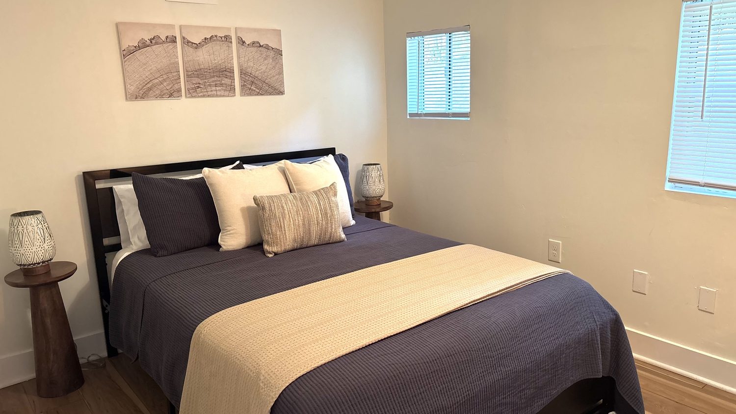 First bedroom — queen bed styled with layered linens and a tan runner, bedside lamps, framed artwork and windows to the woods