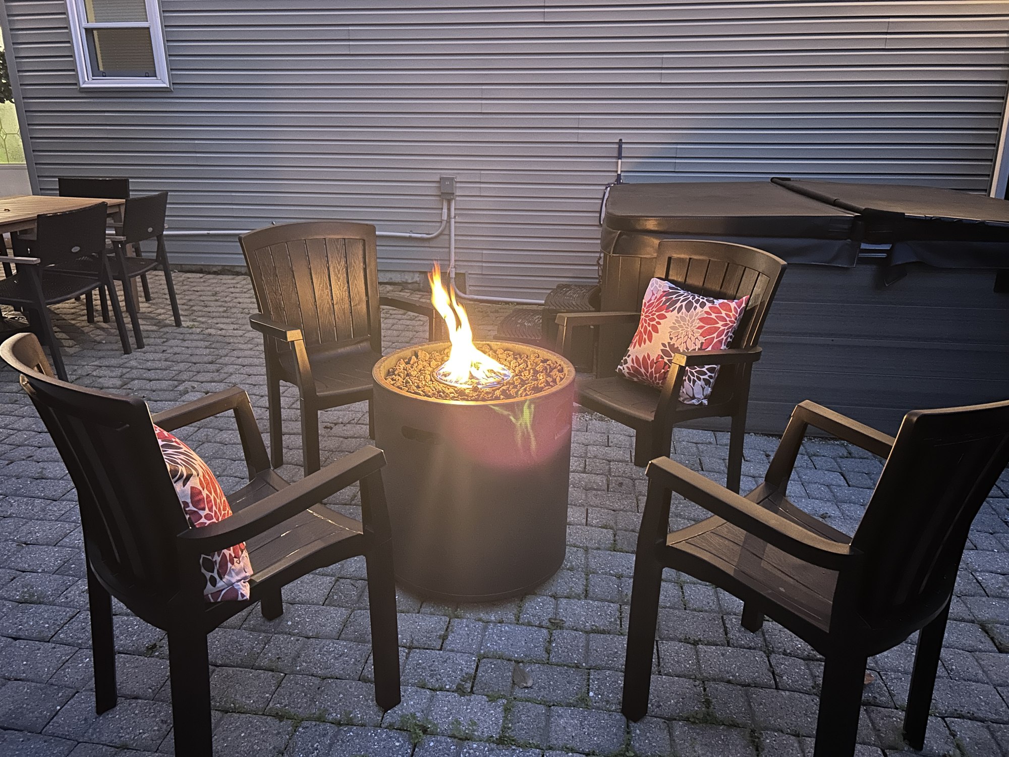 The gas fire pit lit at dusk, four chairs pulled around it with cushions, hot tub alongside
