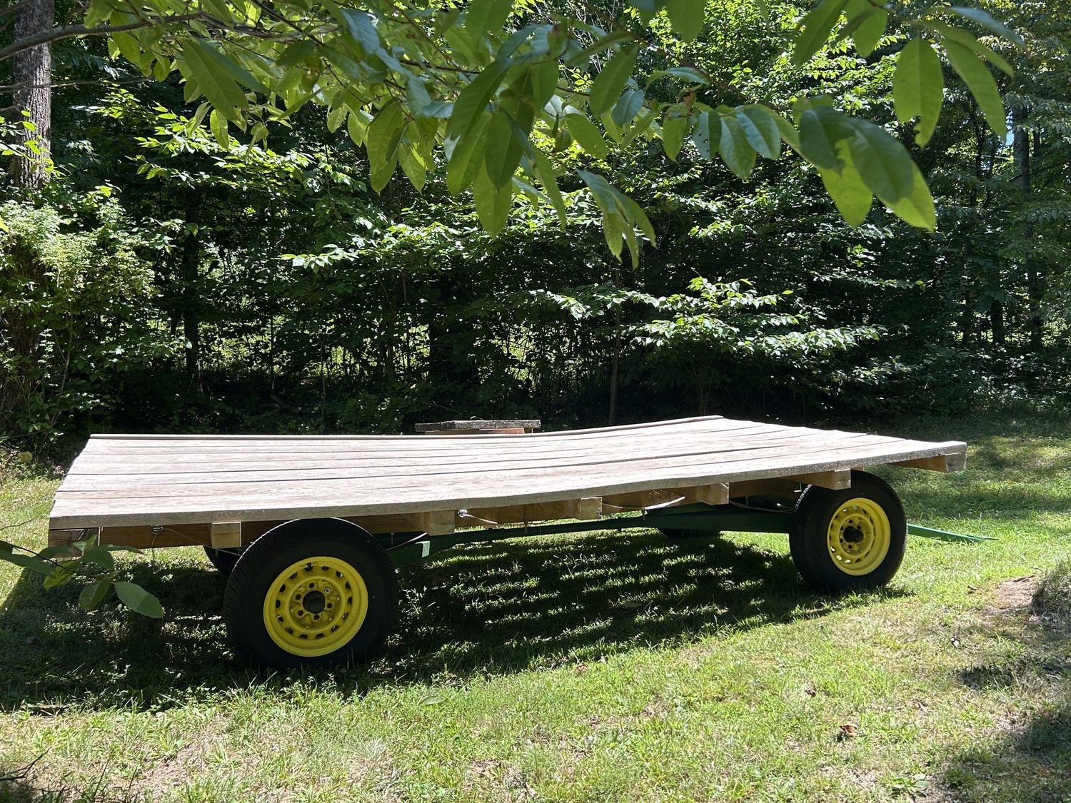 Restored 1965 hay wagon with a weathered plank deck, green frame and yellow wheels, parked on the lawn under the trees