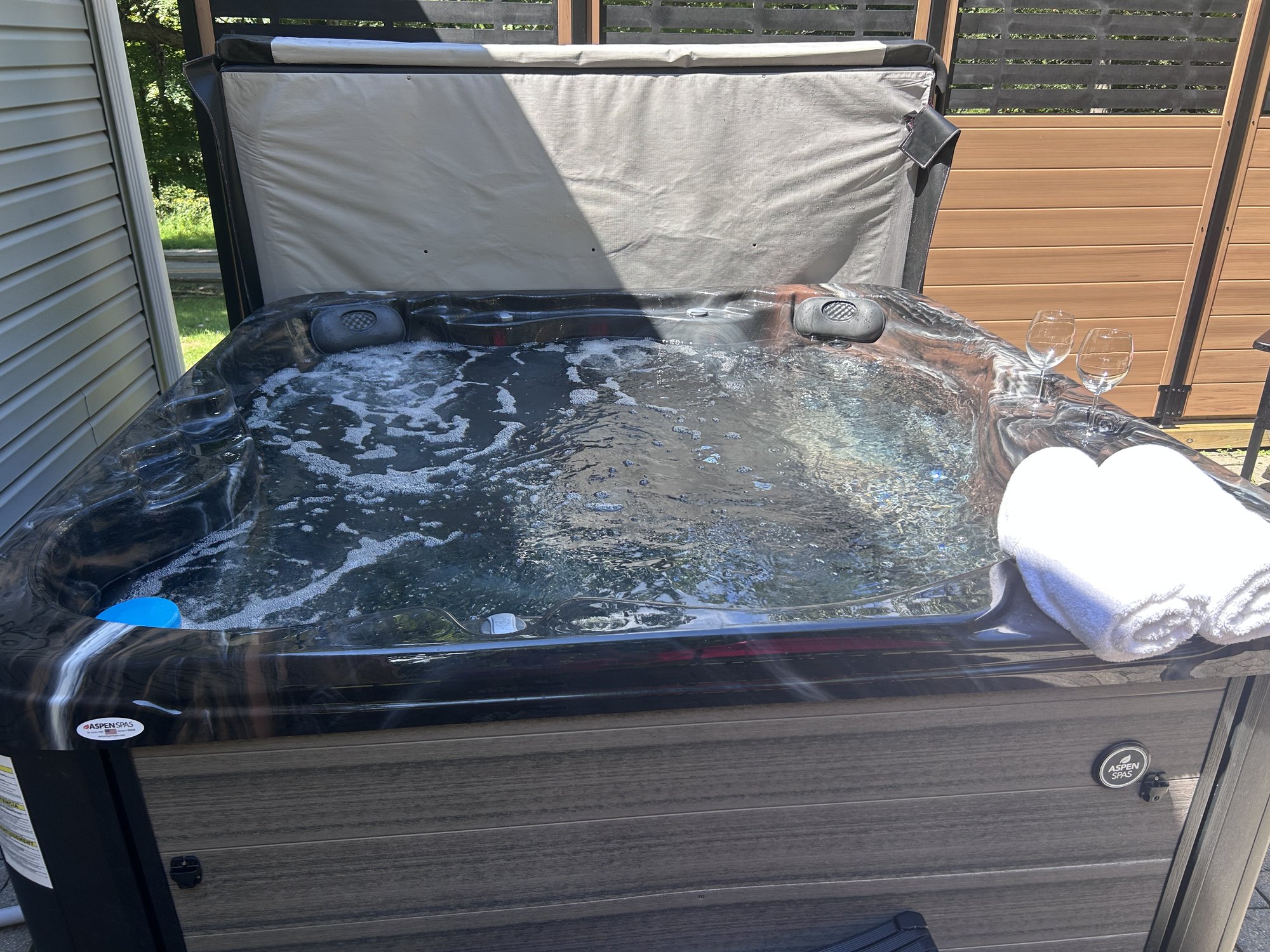Hot tub uncovered and filled, rolled towels and two glasses on the edge, privacy screen behind